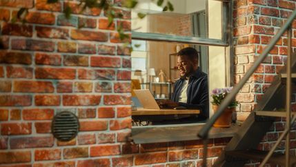 Inside apartment window view of young man smiling and answering emails from home